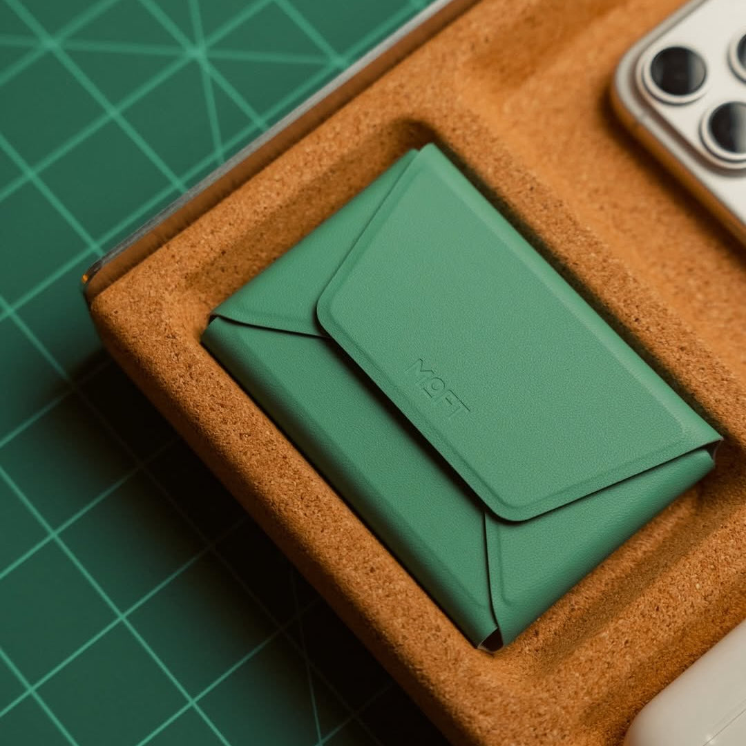 A green envelope-style card holder or wallet with a debossed logo, nestled in a brown textured tray on a green cutting mat, with parts of a phone and a white object visible at the edges.